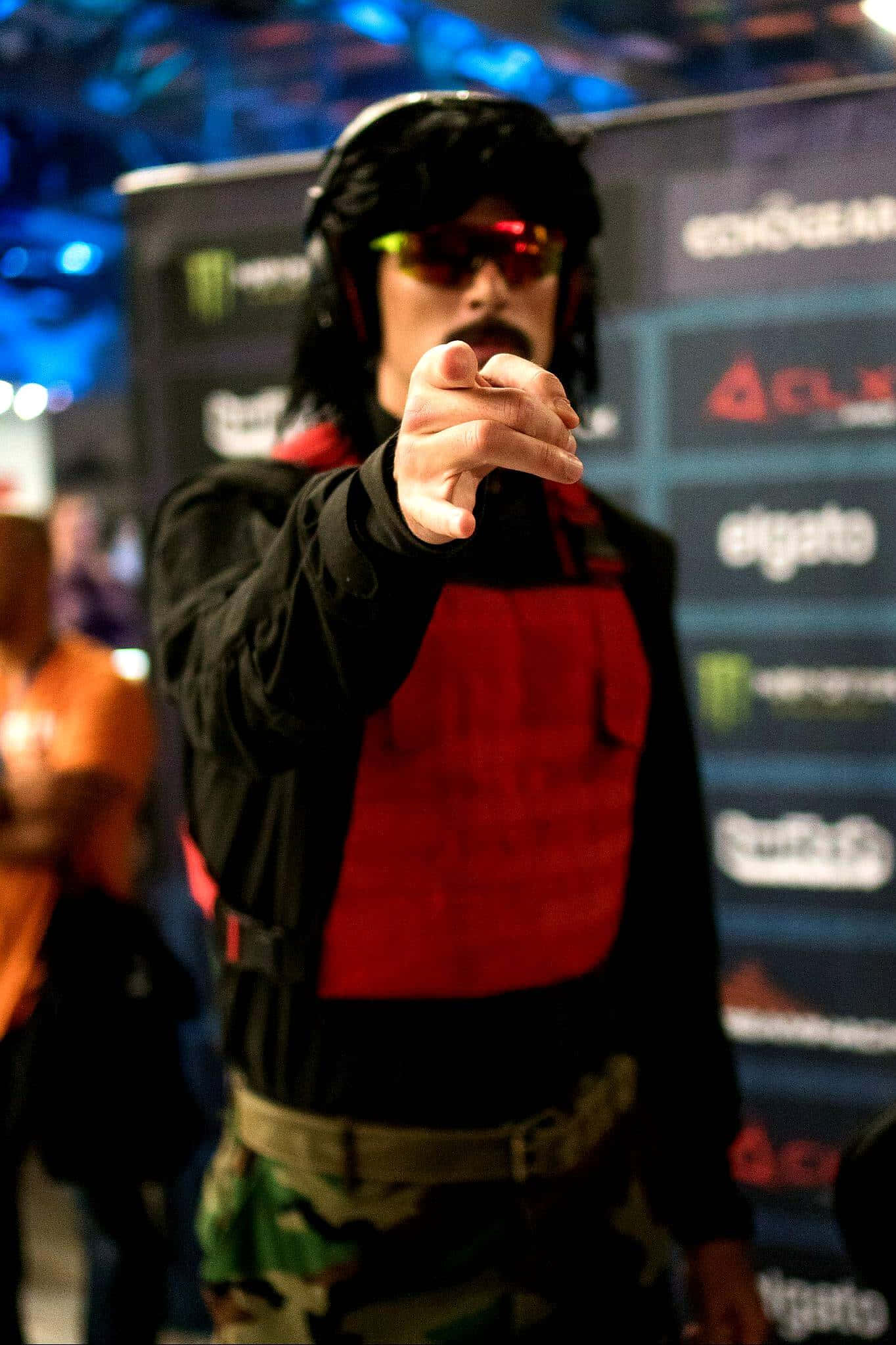 Dr Disrespect 桌布 壁紙 — 一名身穿紅色背心和迷彩褲的男子在藍色舞台燈光下於電子競技活動中直接指向觀眾。