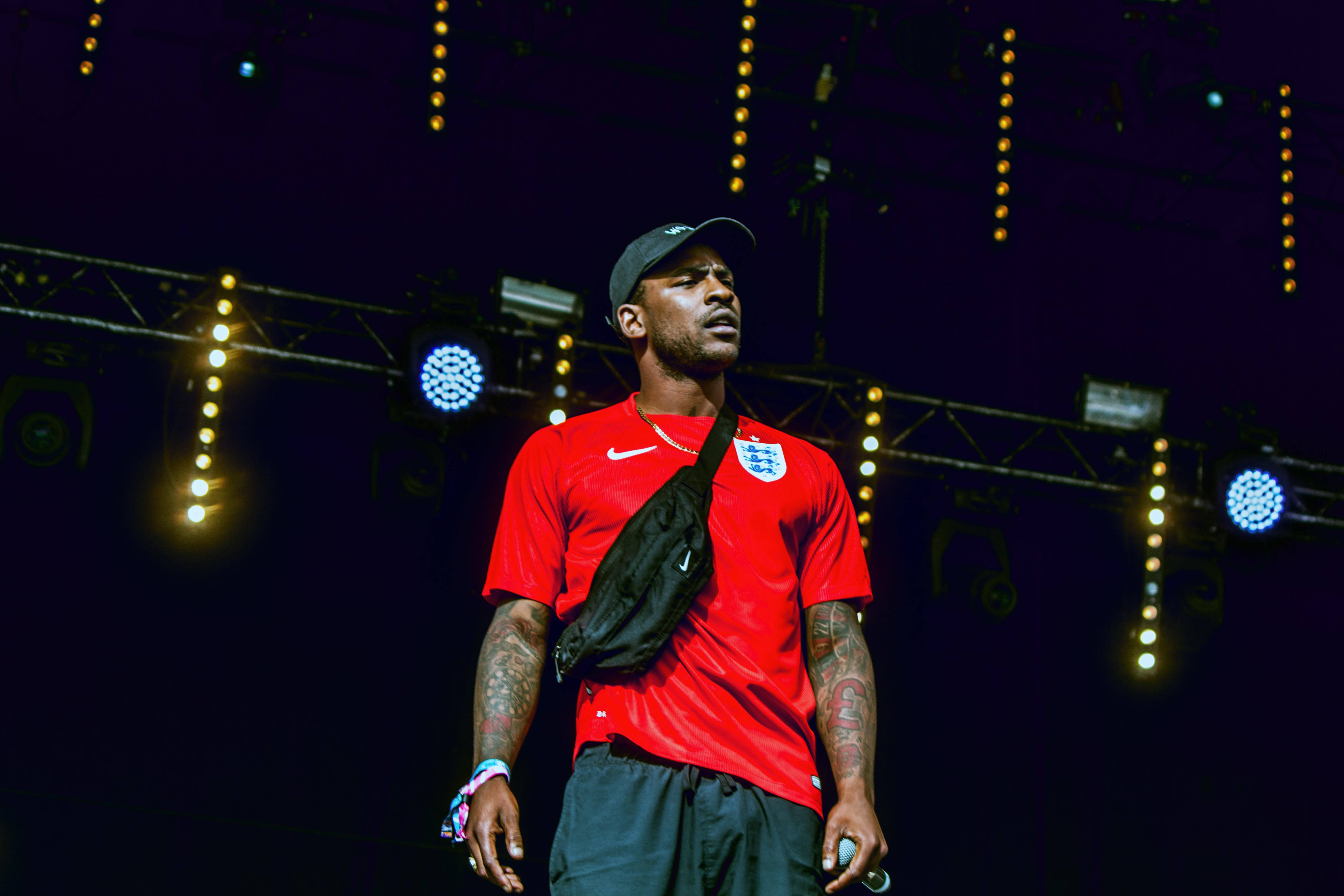Skepta 桌布 壁紙 — 一名身穿紅色英格蘭足球球衣的男子站在燈光昏暗的舞台上，下方有發光的聚光燈和燈串。
