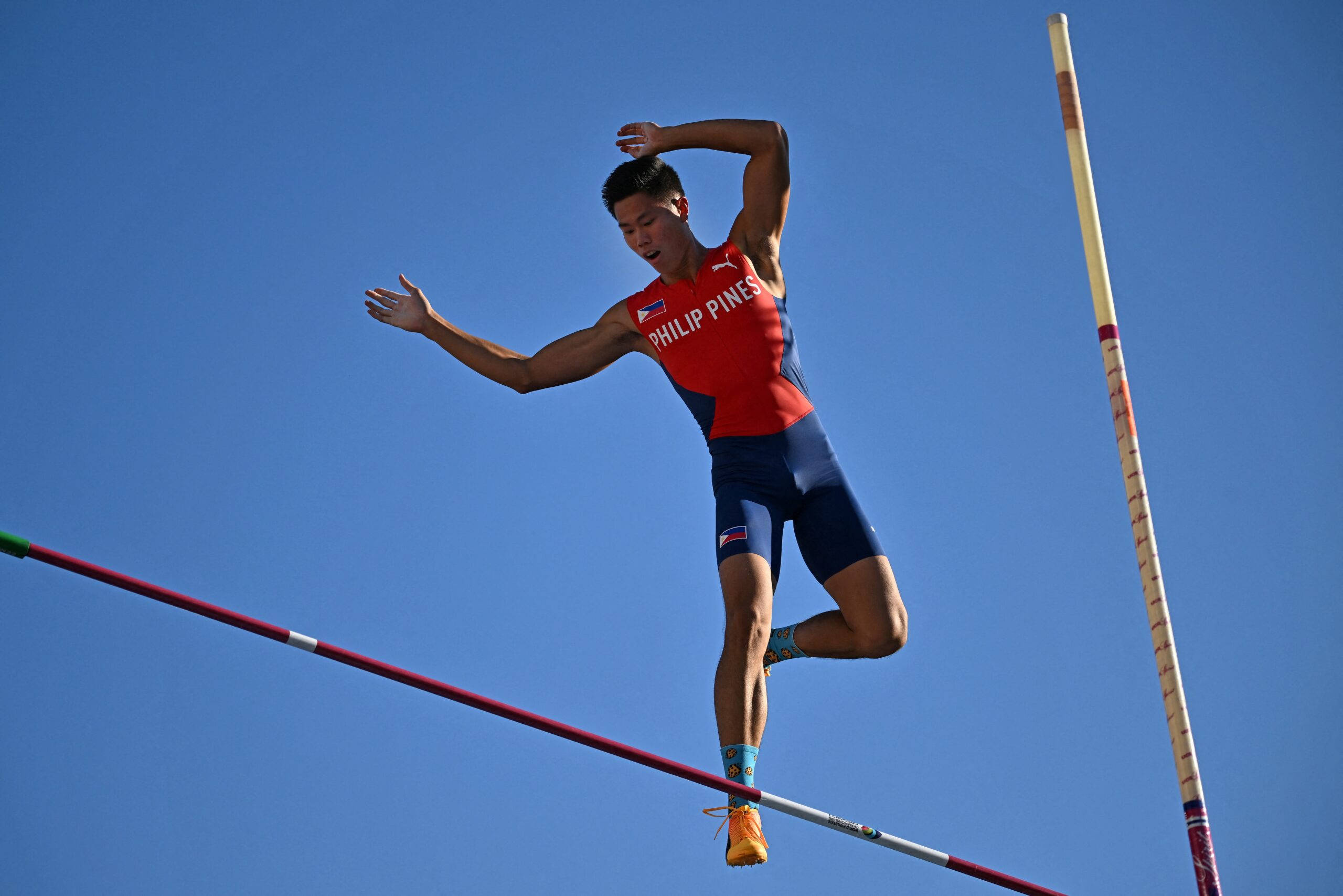 Athletic 桌布 壁紙 — 一名身穿紅色菲律賓制服的男子撐竿跳選手在晴朗藍天下越過橫桿。