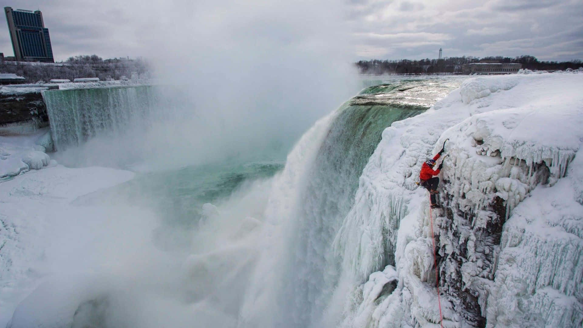 Ice Climbing 桌布 壁紙 — 一名身穿紅色衣服的攀岩者在多雲的天空下，攀爬在咆哮的冰瀑旁邊的冰凍瀑布懸崖上。