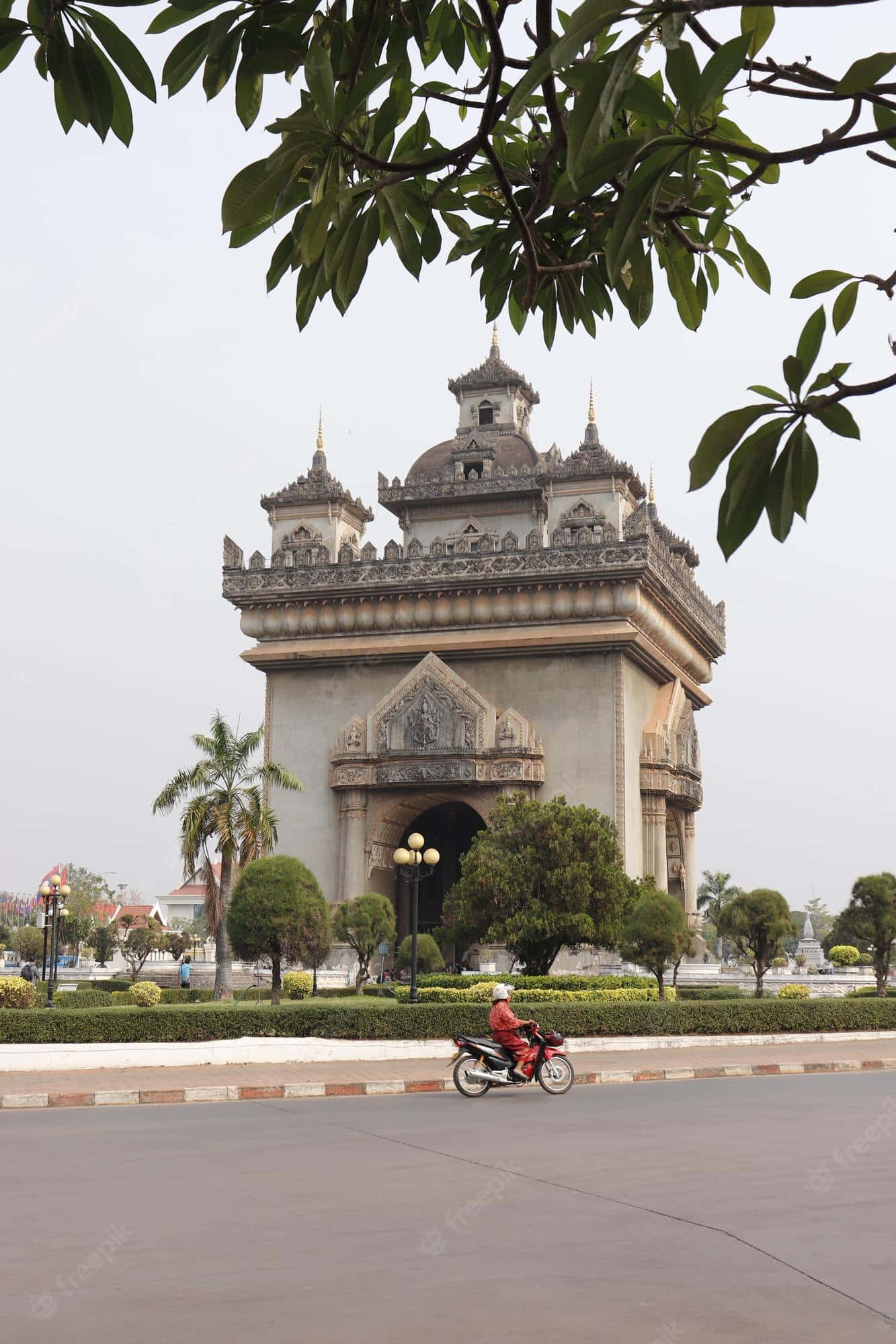 Laos 桌布 壁紙 — 一名身穿紅色衣服的機車騎士穿過一個宏偉華麗的拱門，拱門上方被茂密的綠葉環繞。