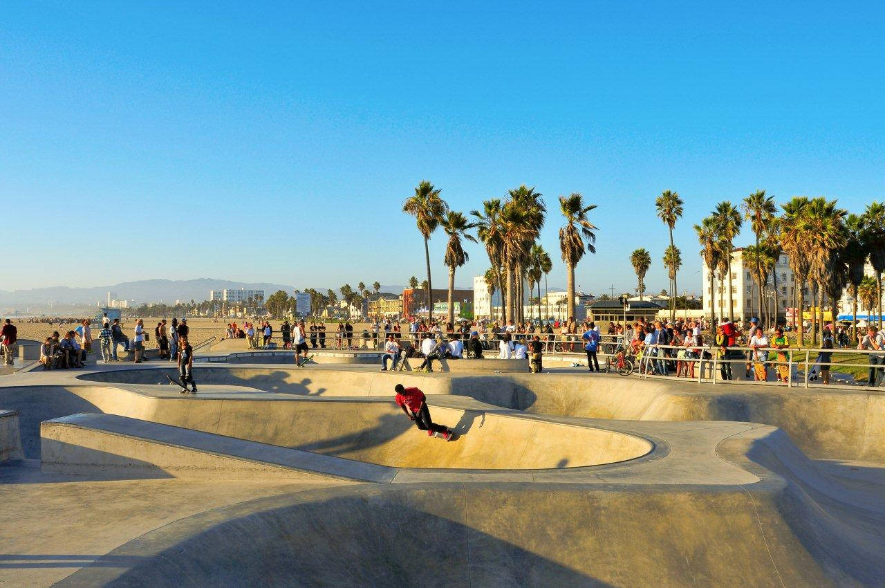 Venice Beach 桌布 壁紙 — 一名身穿紅色衣服的滑板手在陽光明媚的混凝土滑板公園中表演空中技巧，背景有棕櫚樹和海灘遊客。