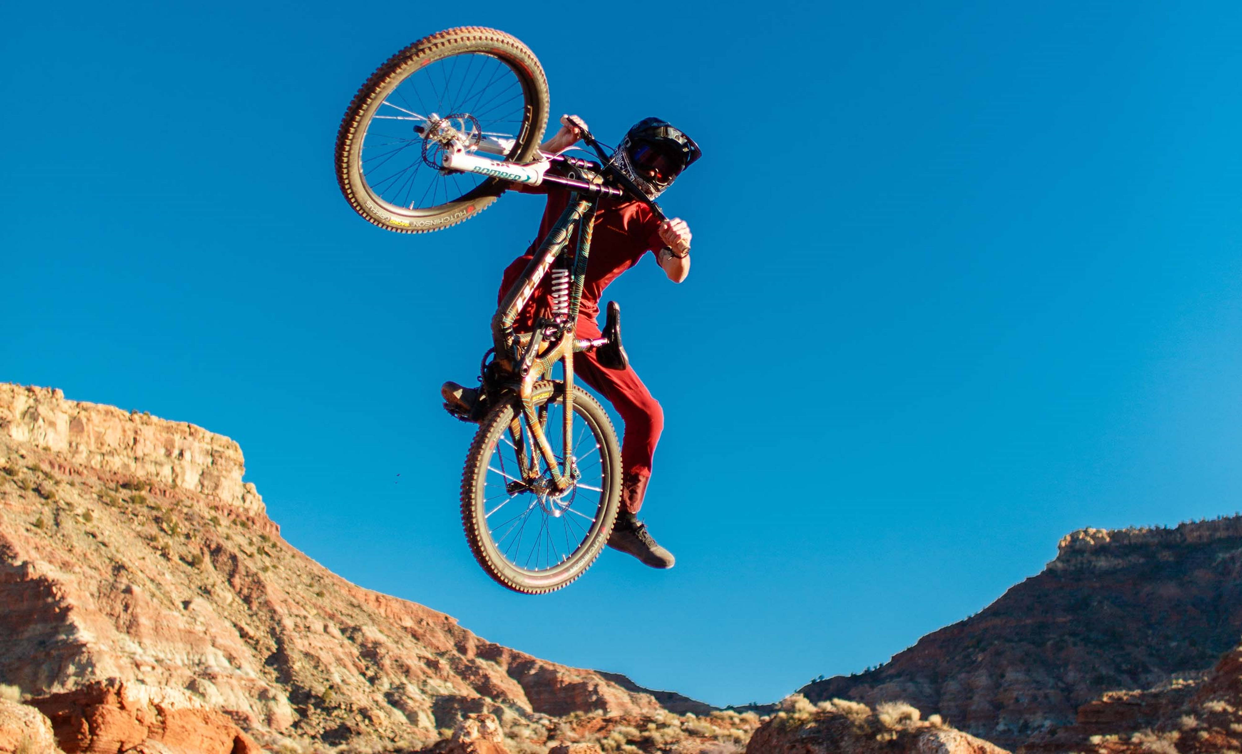 Mountain Bike 桌布 壁紙 — 一名身穿紅色裝備的山地車手在晴朗的藍天下，於紅色岩層上方做出大膽的後空翻。