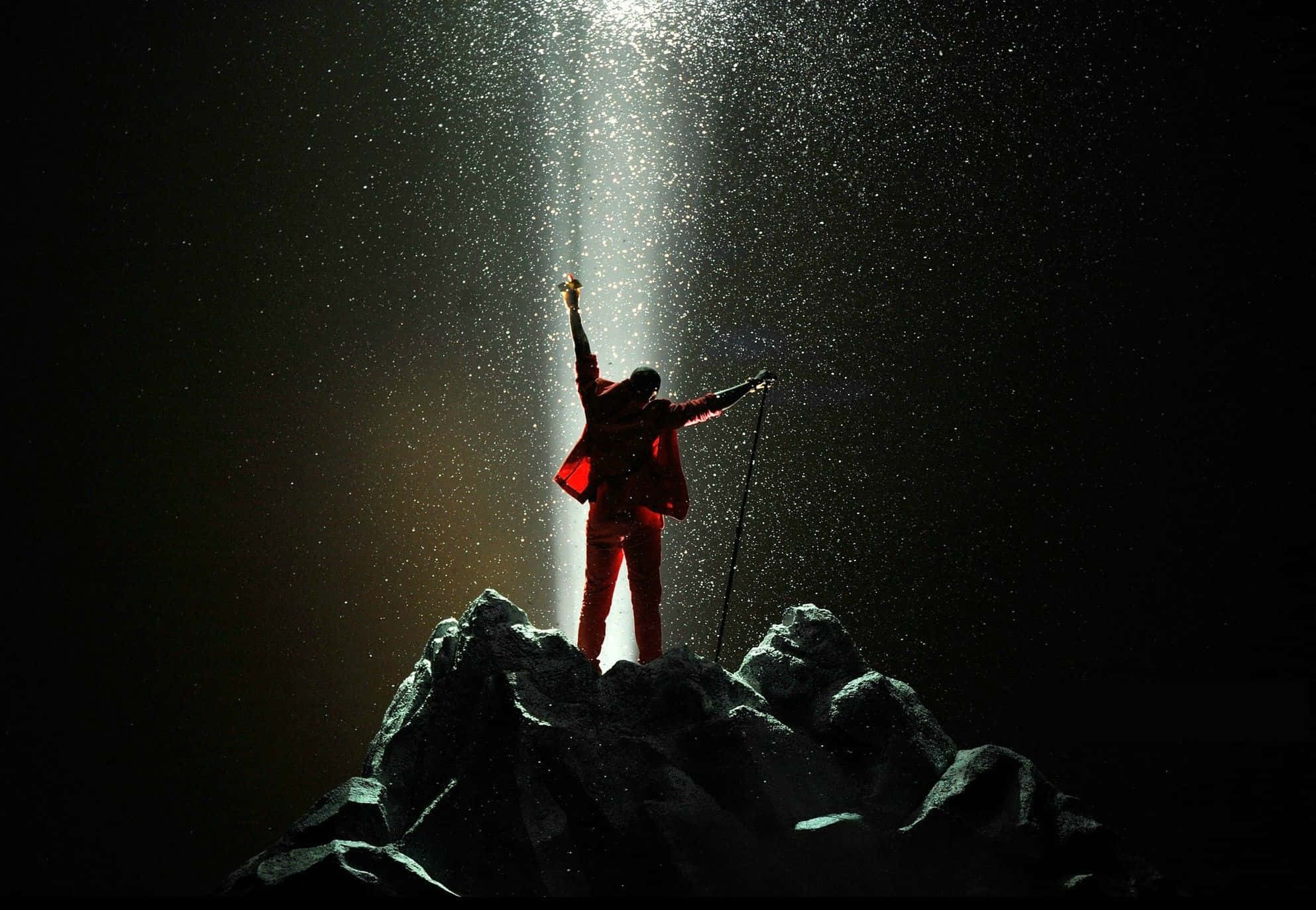 Kanye West Yeezus 桌布 壁紙 — 一名身穿紅色裝備的攀岩者勝利地站在岩石山頂上，一道光束劃破上方的黑暗天空。