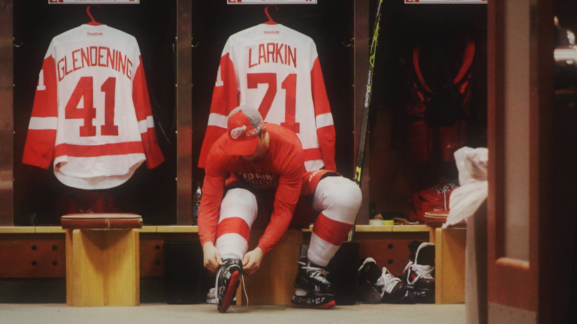 hockey 桌布 壁紙 — 一名身穿紅色裝備的曲棍球運動員坐在更衣室的長凳上繫緊溜冰鞋，身後掛著球衣。