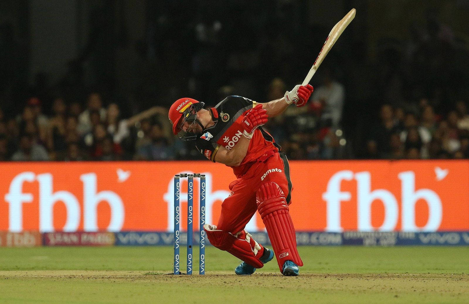royal challengers bangalore 桌布 壁紙 — 一名身穿紅色裝備的板球擊球手在球場燈光下強力揮擊球，背景有模糊的觀眾。