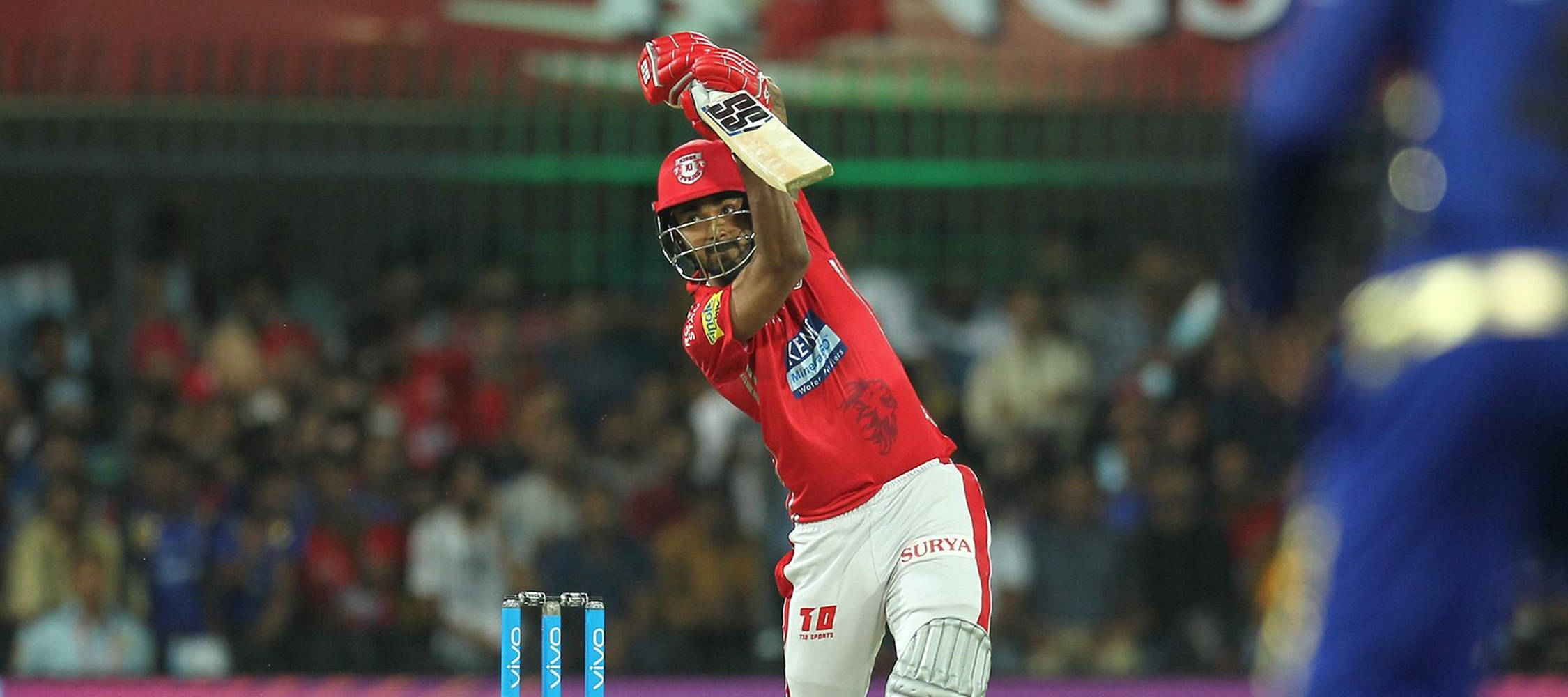 kl rahul 桌布 壁紙 — 一名身穿紅色裝備的板球擊球手在體育場燈光下慶祝強力擊球，背景有觀眾。