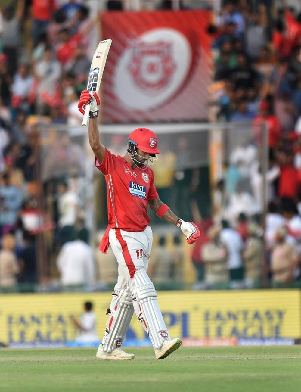 kl rahul 桌布 壁紙 — 一名身穿紅色裝備的板球運動員勝利地高舉球棒，球迷在體育場內歡呼。