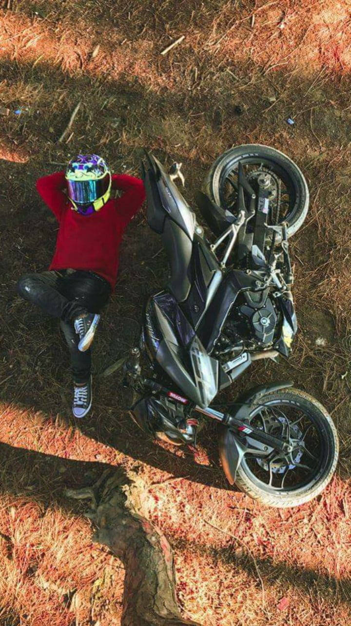 機車 桌布 壁紙 — 一名身穿紅色襯衫和彩色頭盔的摩托車騎士，在斑駁的陽光下，躺在乾草上，旁邊有一輛倒下的黑色摩托車。
