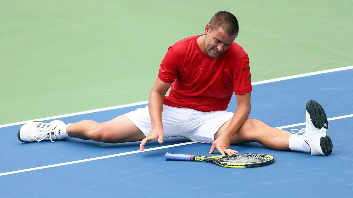 Tennis 桌布 壁紙 — 一名身穿紅色襯衫和白色短褲的男網球運動員坐在藍色球場上，痛苦地抱著腿，球拍放在旁邊。