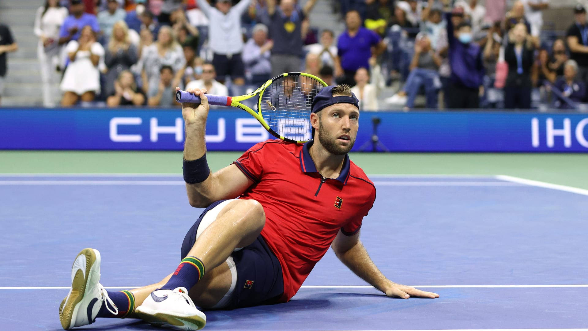 Tennis 桌布 壁紙 — 一名身穿紅色襯衫和藍色短褲的男網球運動員在打出致勝球後，握著球拍倒在藍色球場上。