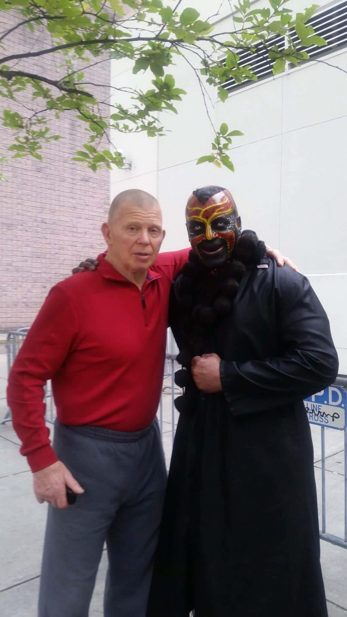 WWE 桌布 壁紙 — 一名身穿紅色襯衫的男子，在枝葉茂密的樹枝下，與一位身穿黑色外套、戴著面具的摔角手合影。
