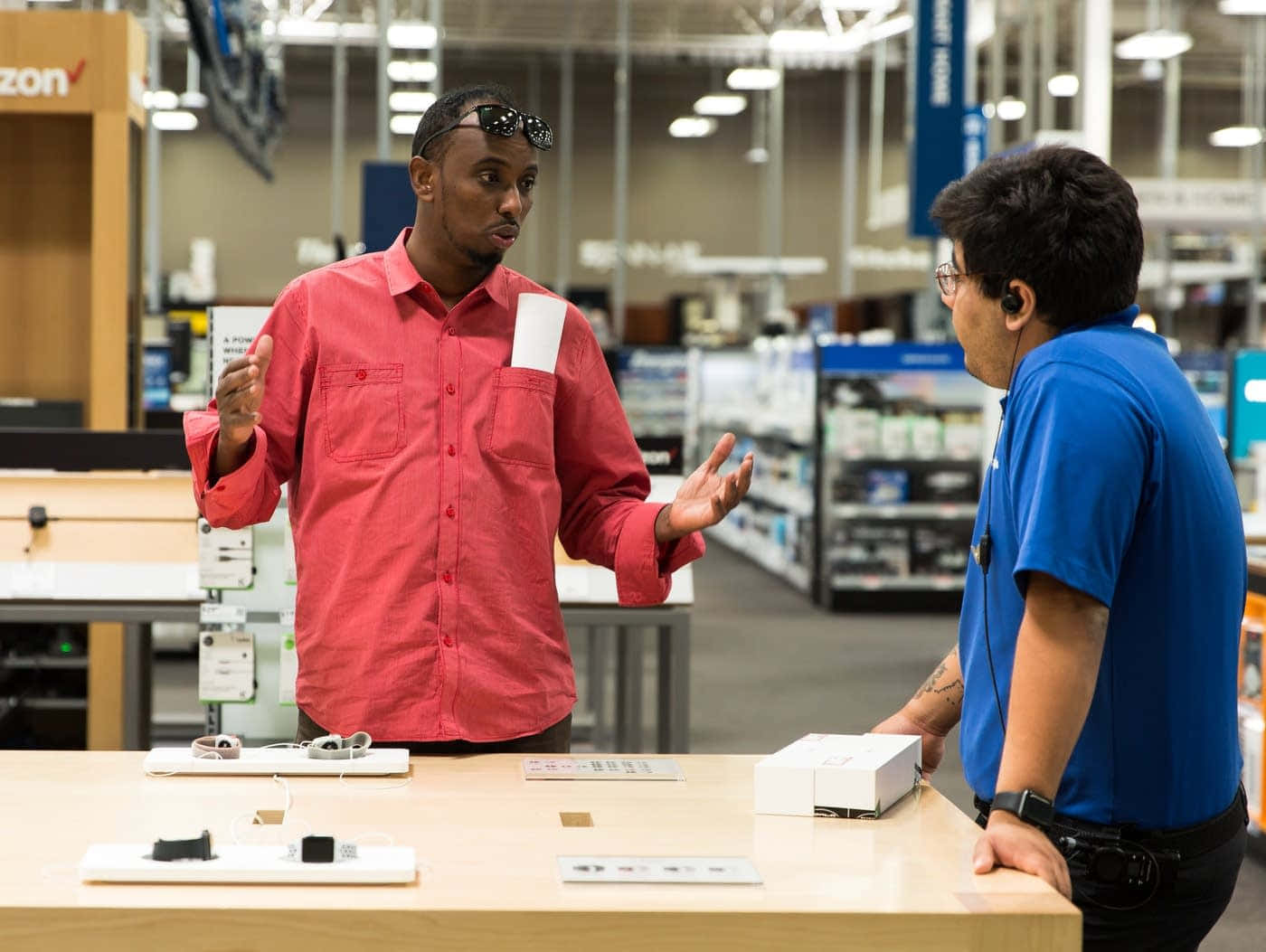 Best Buy 桌布 壁紙 — 一名身穿紅色襯衫的男子在光線明亮的零售店電子產品櫃檯前，與一名身穿藍色襯衫的店員交談，並生動地做著手勢。