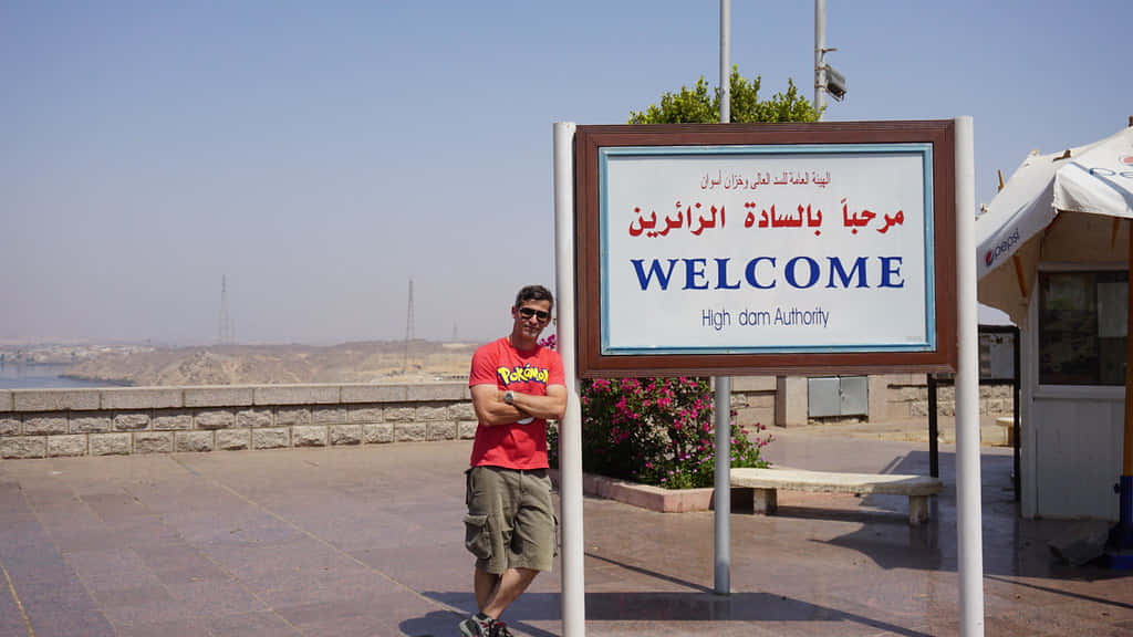 沙漠 桌布 壁紙 — 一名身穿紅色襯衫的男子自信地交叉雙臂，站在高壩管理局場地的雙語歡迎標誌旁，背景是晴朗的藍天。