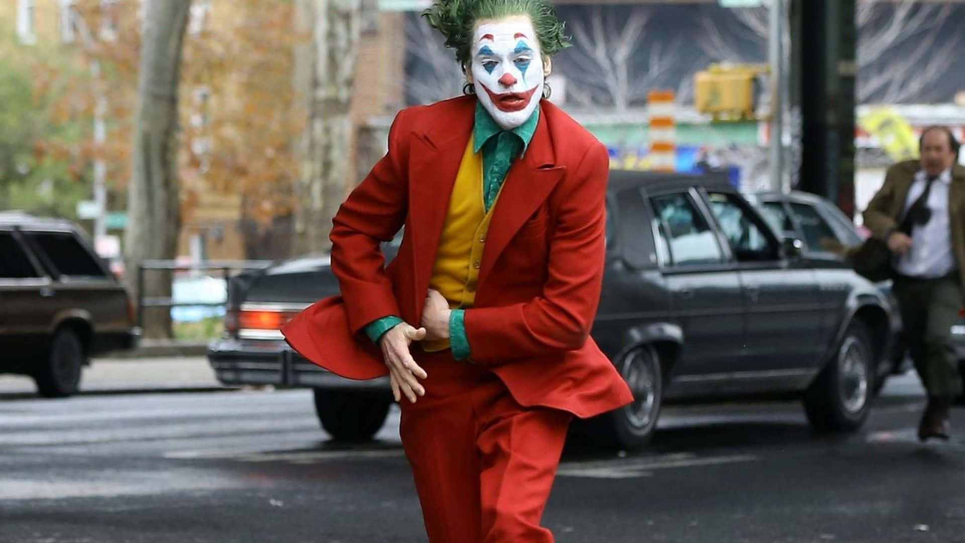 桌布 壁紙 — 一名身穿紅色西裝、化著小丑妝的男子，在背景有汽車和行人的城市街道上充滿活力地跳舞。