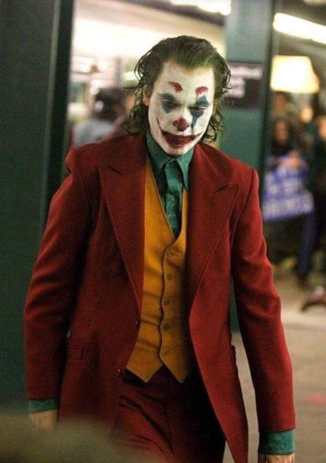 Joker 2019 桌布 壁紙 — 一名身穿紅色西裝、化著小丑妝的男子，帶著陰險的笑容走過燈光昏暗的地鐵站。