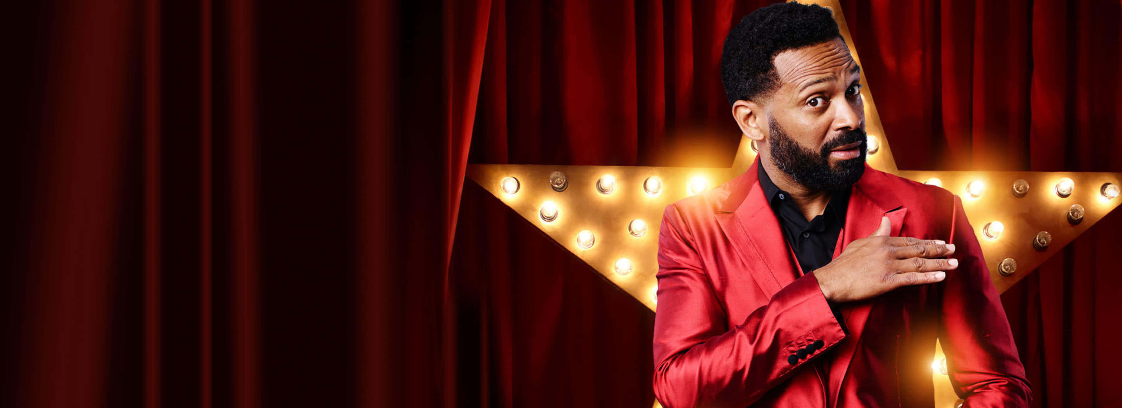 Mike Epps 桌布 壁紙 — 一名身穿紅色西裝的男子自信地站在發光的星星和紅色帷幕前，手放在胸前，彷彿在舞台上表演。