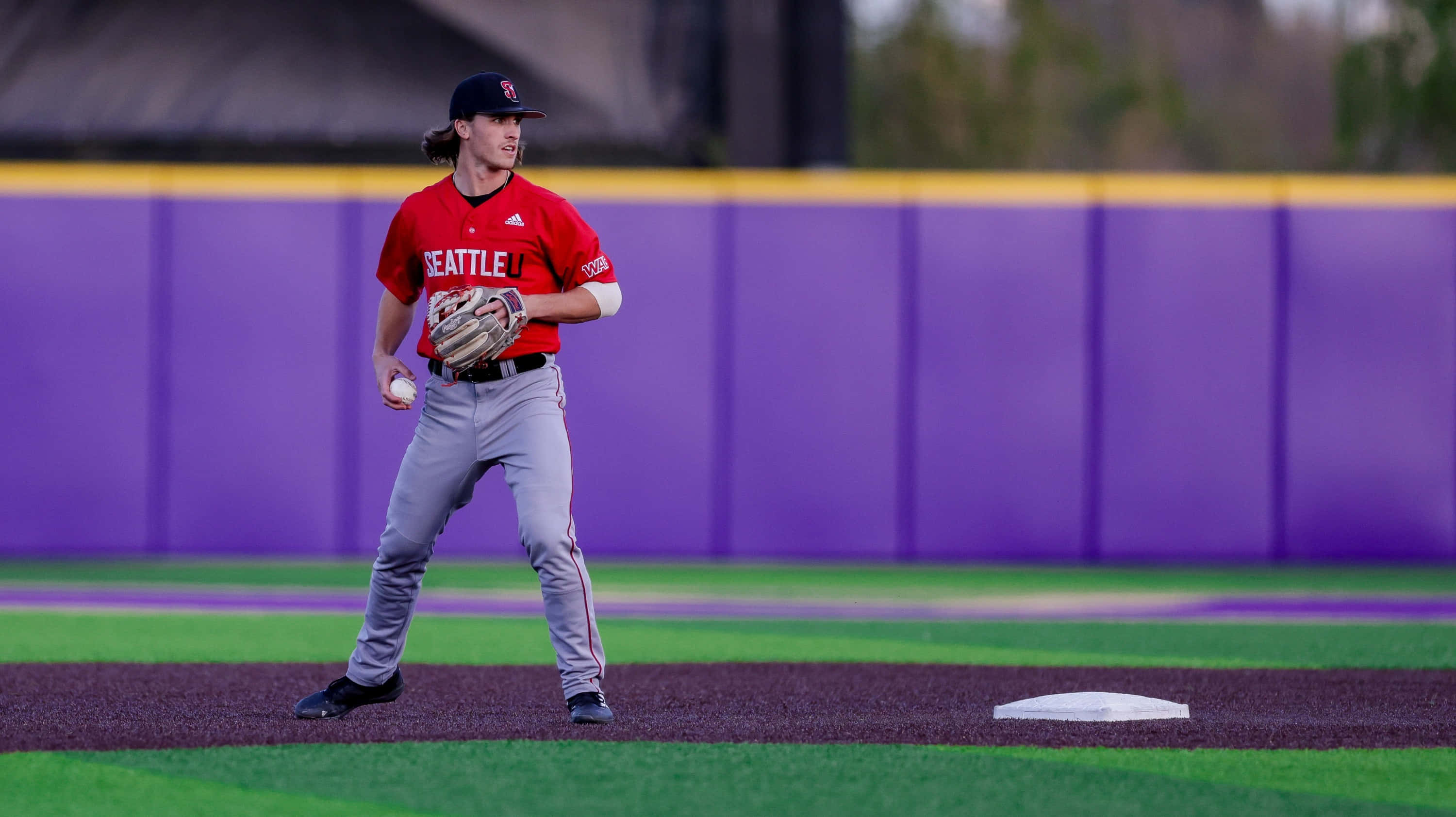 College Baseball 桌布 壁紙 — 一名身穿紅色西雅圖球衣的棒球運動員在紫色牆壁環繞的鮮綠色場地上警覺地站在壘包附近。