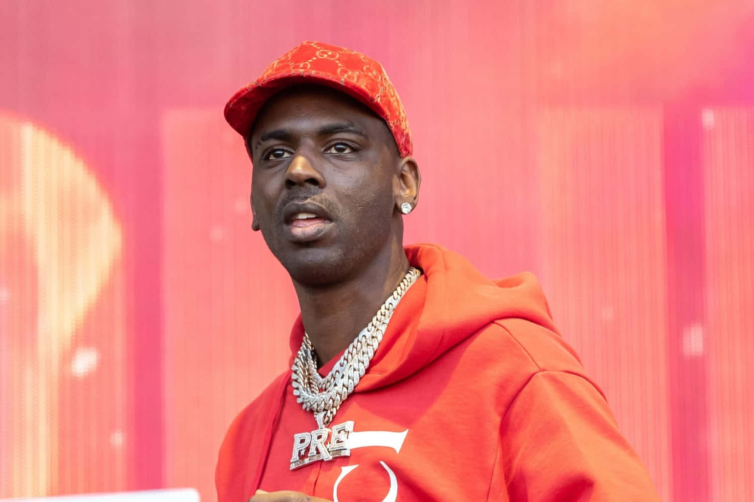 Young Dolph 桌布 壁紙 — 一名身穿紅色連帽衫和帽子的男子站在充滿活力的粉紅色背景前，帶有發光效果。