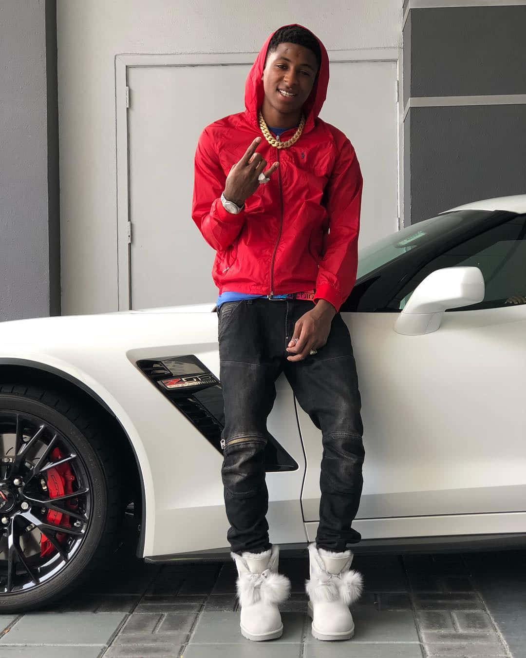 NBA YoungBoy 桌布 壁紙 — 一名身穿紅色連帽衫和白色毛靴的年輕男子在車庫裡自信地站在一輛白色跑車旁邊。