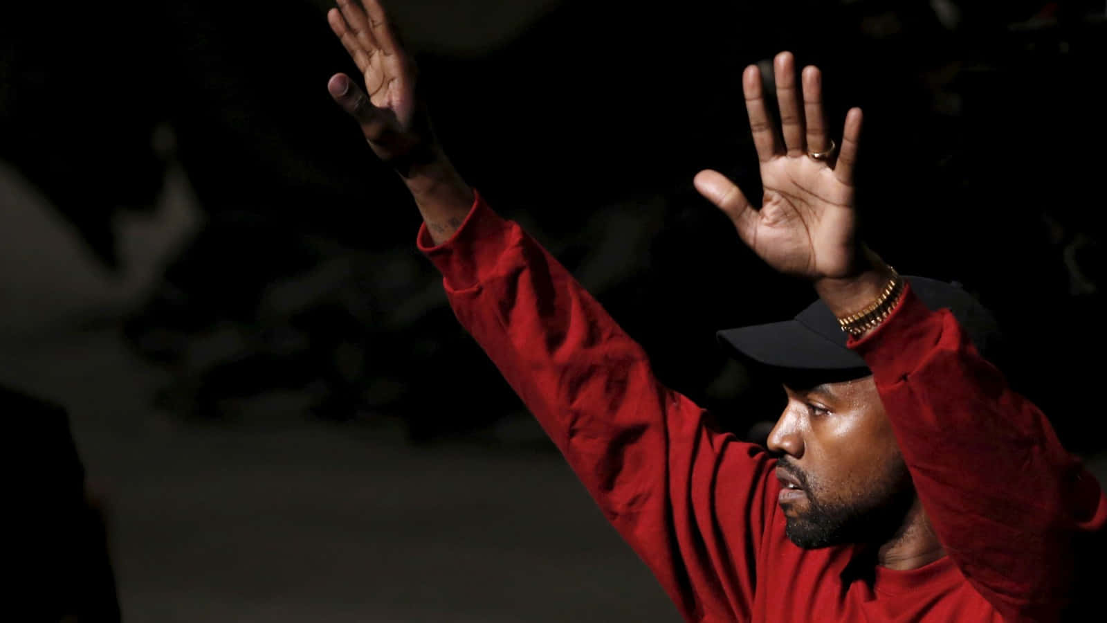 The Life Of Pablo 桌布 壁紙 — 一名身穿紅色運動衫和黑色帽子的男子，在深色背景下舉起雙手，擺出戲劇性的姿勢。