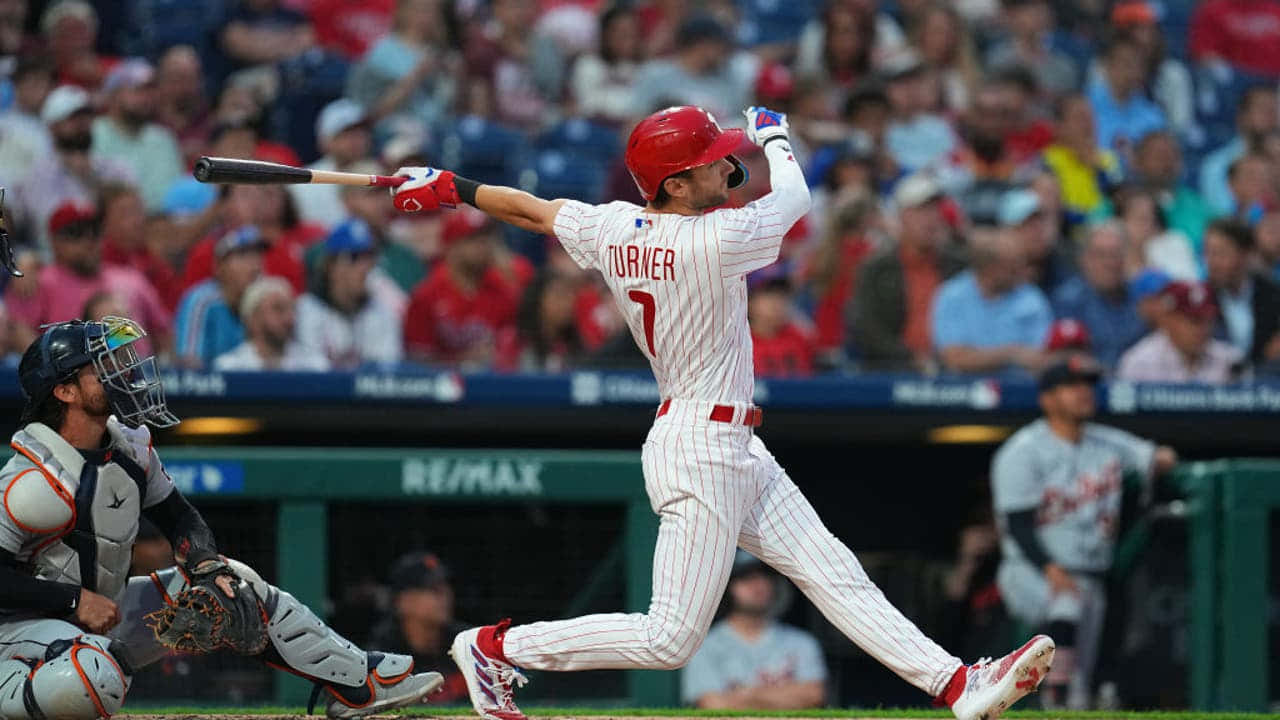 Trea Turner Phillies 桌布 壁紙 — 一名身穿紅色頭盔和白色細條紋球衣的棒球選手，在日間比賽中全力揮棒，背景有捕手和觀眾。