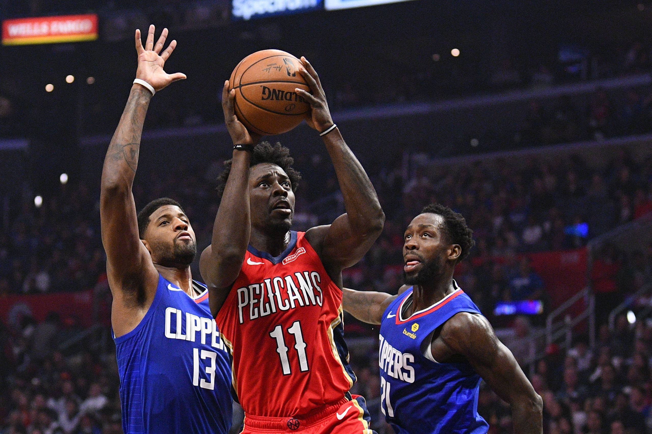 Jrue Holiday 桌布 壁紙 — 一名身穿紅色鵜鶘隊球衣的籃球運動員在場館燈光下跳投，兩名快艇隊防守球員試圖封阻該投籃。
