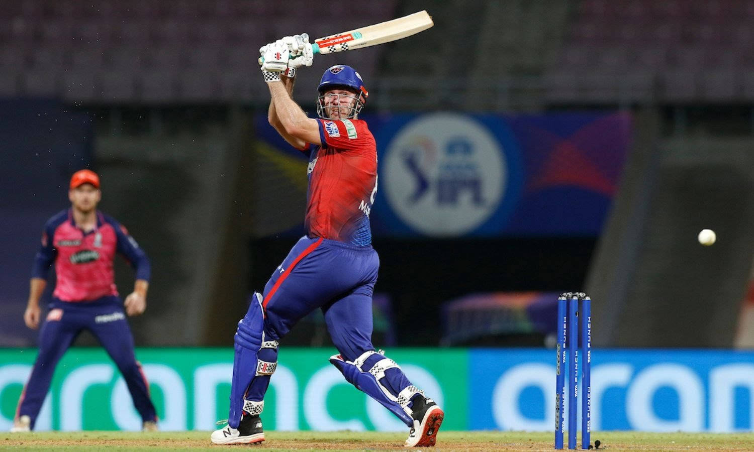 Delhi Capitals 桌布 壁紙 — 一名身穿紅藍球衣的板球擊球手在夜間比賽中，在體育場燈光下強力揮棒擊球。