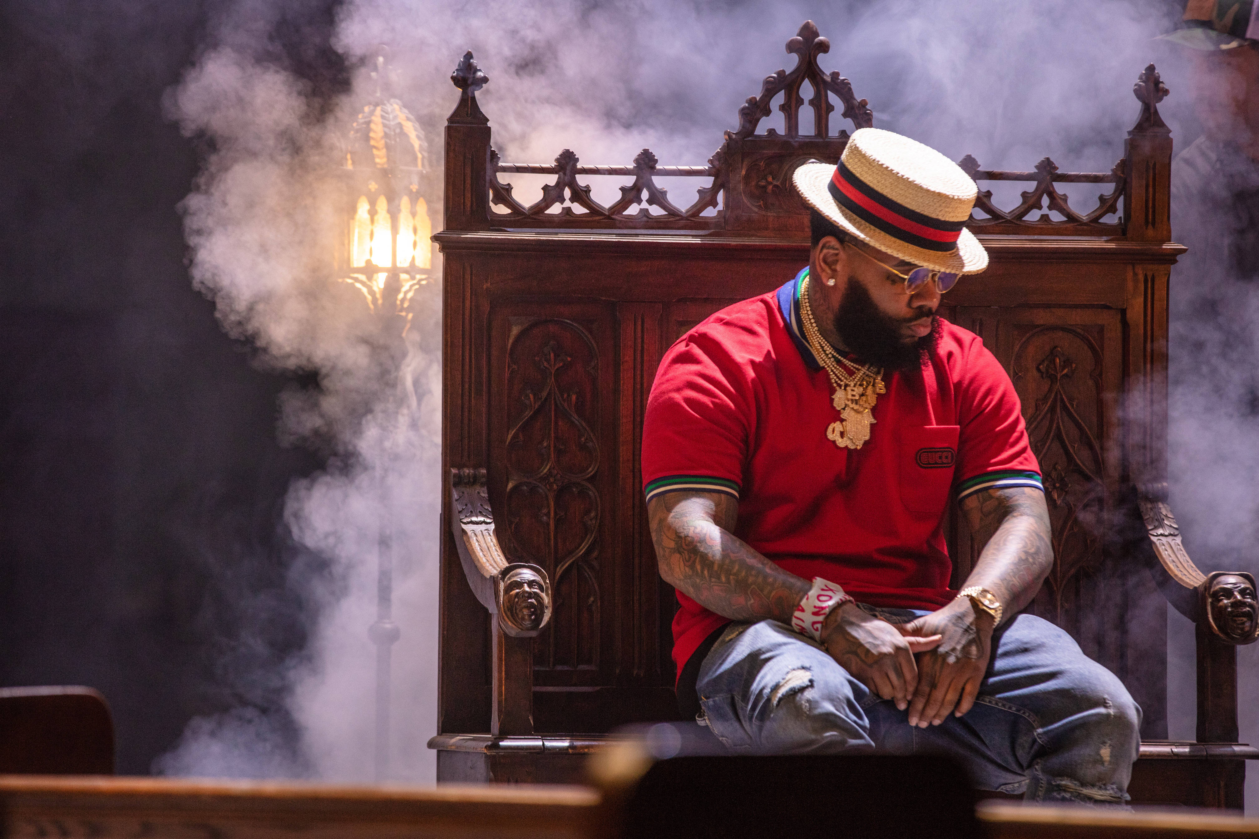 Kevin Gates 桌布 壁紙 — 一名身穿紅衫、頭戴草帽的男子，在煙霧繚繞和溫暖燈光中，威嚴地坐在華麗的木製寶座上。