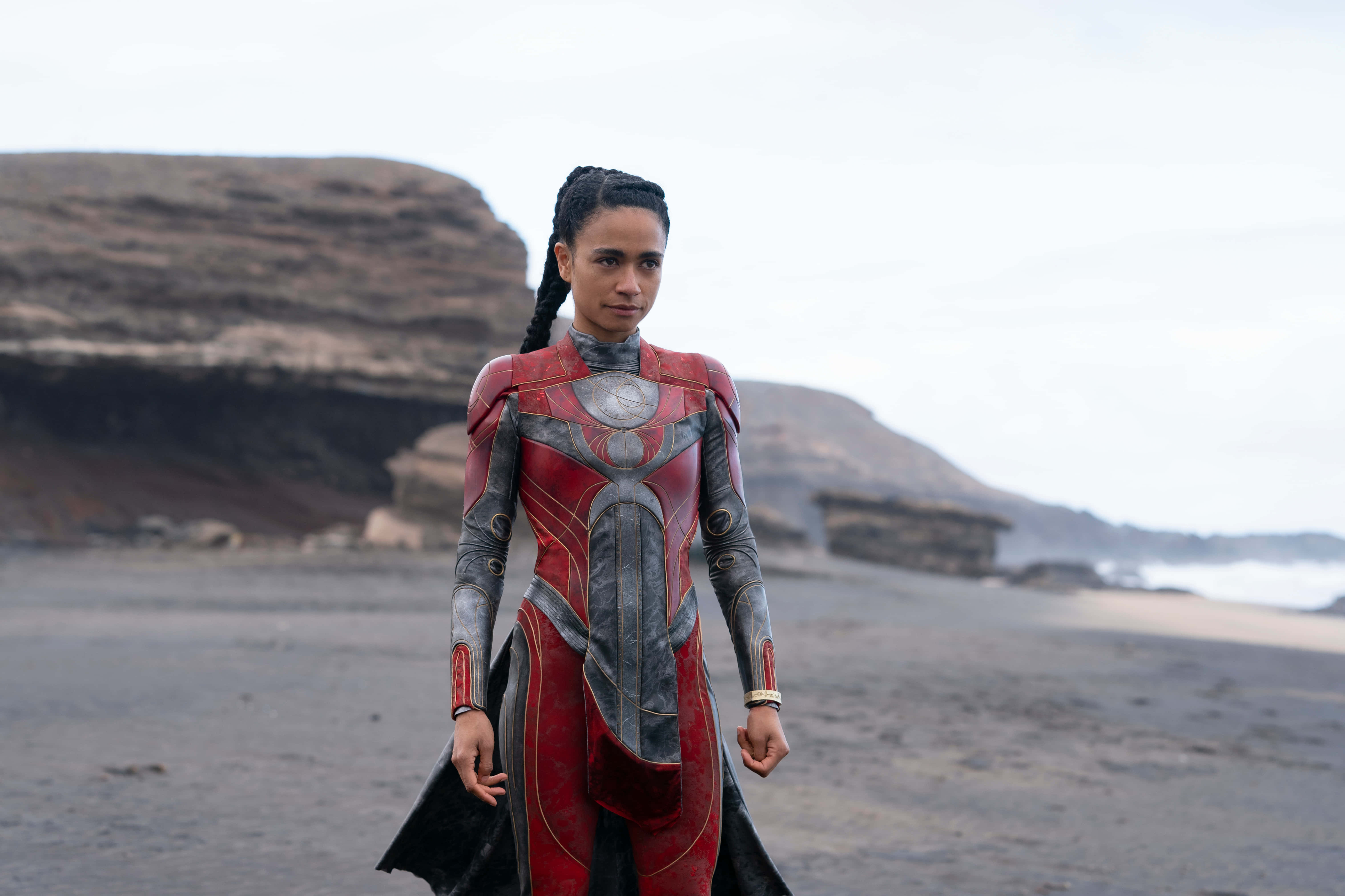 Eternals Movie 桌布 壁紙 — 一名身穿紅銀色盔甲套裝的女性自信地站在黑暗的火山海灘上，身後是層疊的懸崖。
