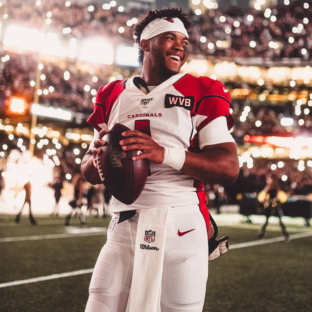 運動 桌布 壁紙 — 一名身穿紅雀隊裝備、面帶微笑的NFL四分衛在燈火通明的球場上抱著橄欖球，背景是歡呼的球迷。
