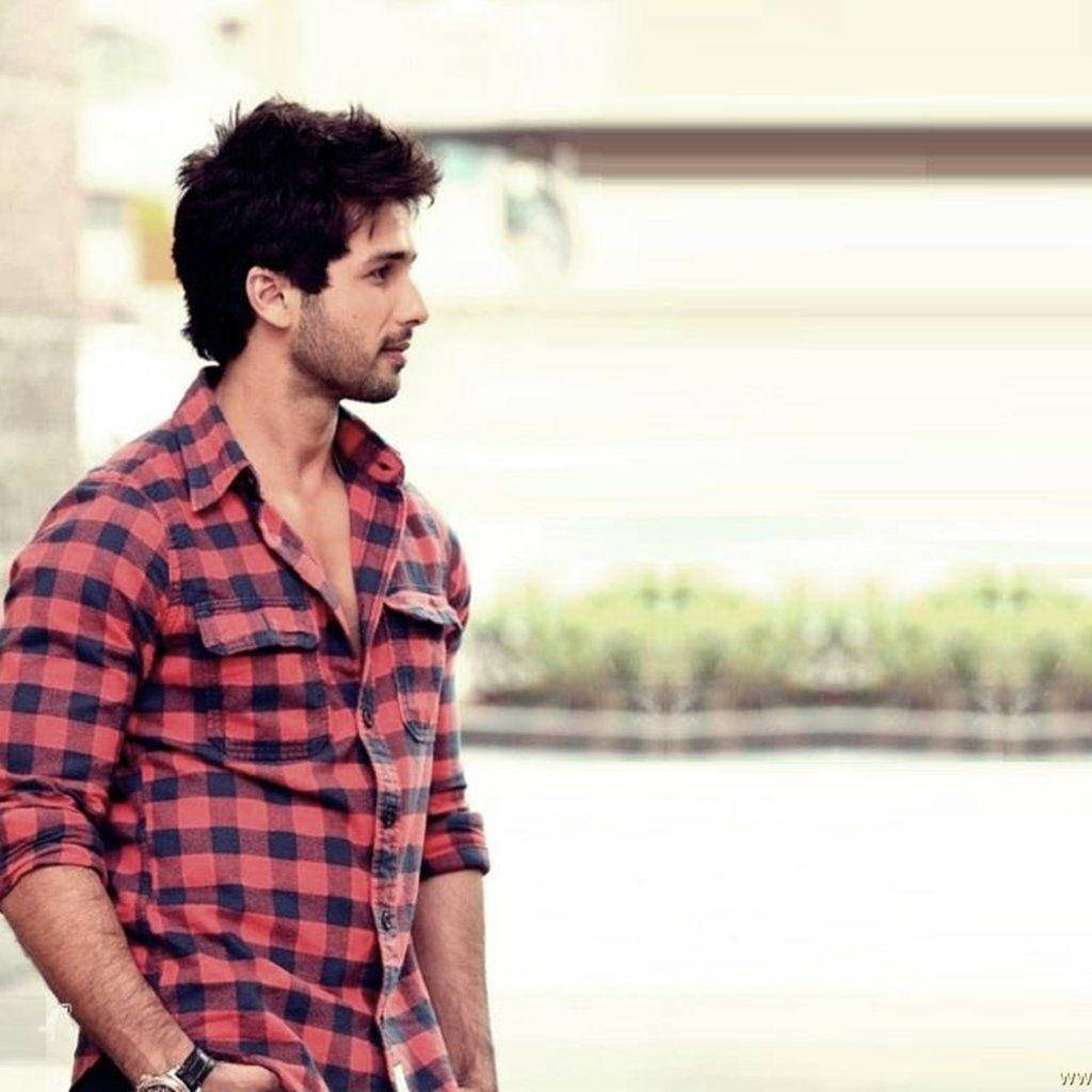 Shahid Kapoor 桌布 壁紙 — 一名身穿紅黑格子襯衫的男子，在模糊的城市背景下，若有所思地站在水邊。