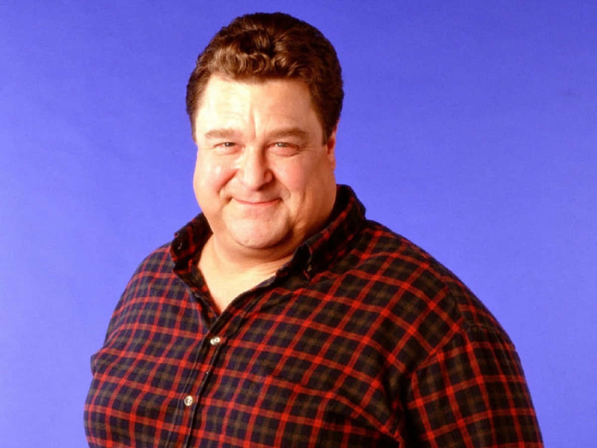 John Goodman 桌布 壁紙 — 一名身穿紅黑格紋襯衫的男士在純藍色攝影棚背景前溫暖地微笑。