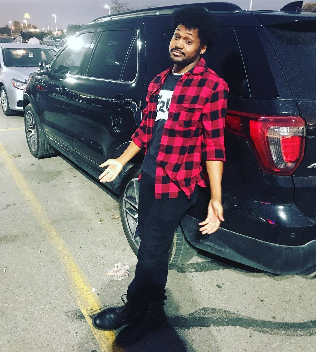 Coryxkenshin 桌布 壁紙 — 一名身穿紅黑格紋襯衫的男子在夜晚燈光昏暗的停車場中，隨意地靠在一輛黑色SUV上。