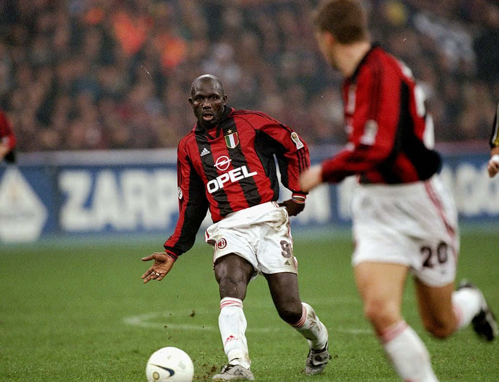 Ac Milan 桌布 壁紙 — 一名身穿紅黑條紋的足球員在綠色球場上運著白色足球，背景有模糊的觀眾。