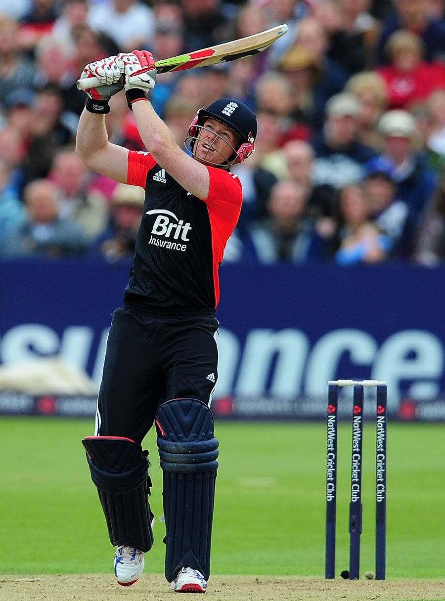 eoin morgan 桌布 壁紙 — 一名身穿紅黑球衣的板球擊球手在比賽中強力揮動球棒，背景有觀眾。