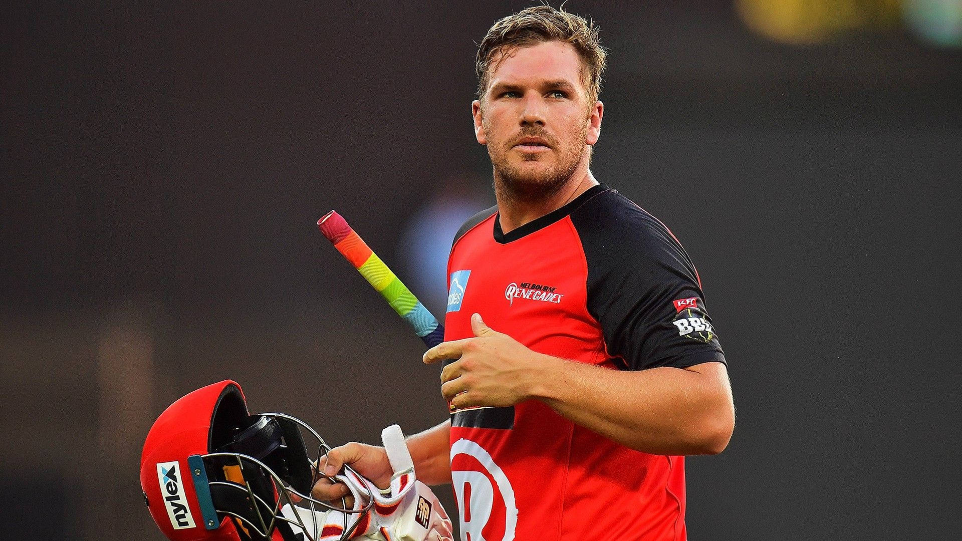 Aaron Finch 桌布 壁紙 — 一名身穿紅黑色球衣的板球運動員在模糊的體育場場地上自信地做手勢，手持球棒和頭盔。