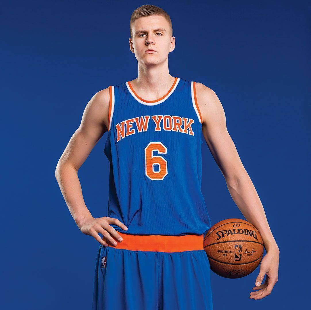 Kristaps Porzingis 桌布 壁紙 — 一名身穿紐約尼克隊球衣的籃球運動員自信地站在純藍色攝影棚背景前，手持一個Spalding籃球。