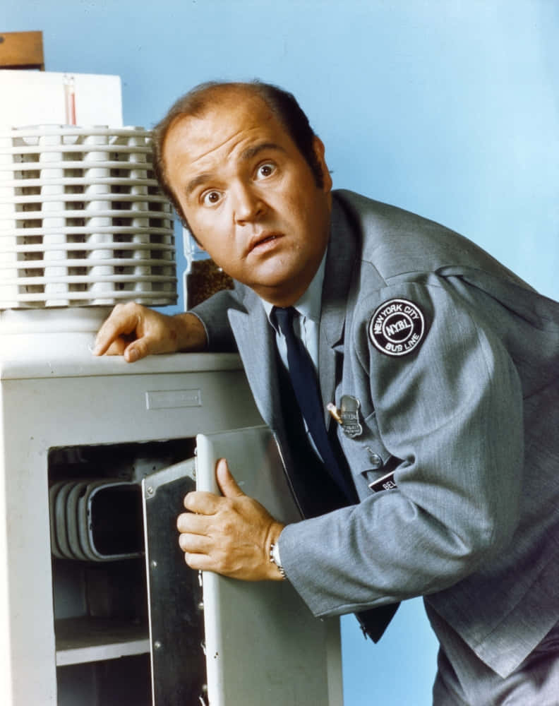 Dom Deluise 桌布 壁紙 — 一名身穿紐約市巴士司機制服的男人，在素淨的藍色牆壁前，從巴士引擎艙後面睜大眼睛窺視。