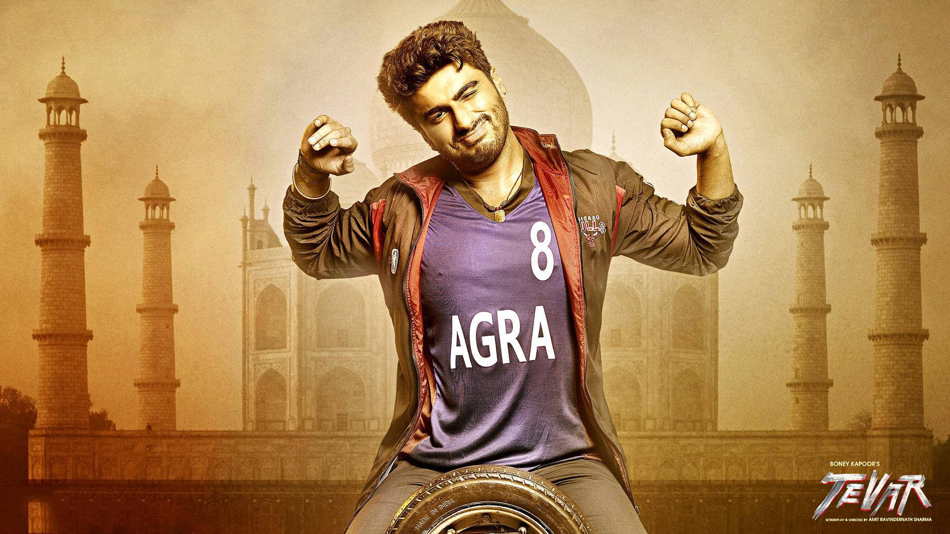 Arjun Kapoor 桌布 壁紙 — 一名身穿紫色 'AGRA' 球衣的男子在朦朧的泰姬瑪哈陵背景下勝利地展示肌肉。