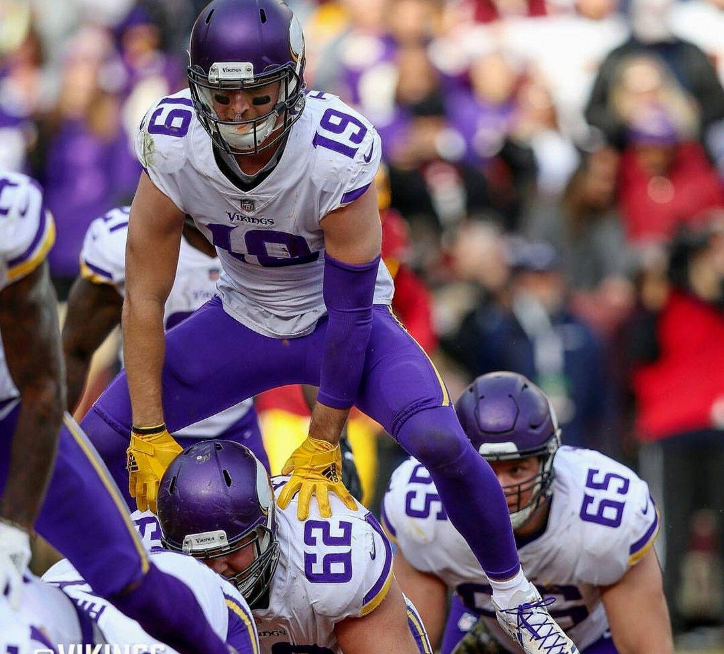 Adam Thielen 桌布 壁紙 — 一名身穿紫色和白色裝備的明尼蘇達維京人美式足球員在比賽中跳過隊友，背景是模糊的人群。