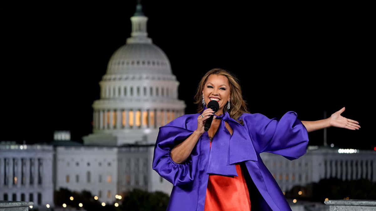 Vanessa Williams 桌布 壁紙 — 一名身穿紫色和紅色長裙的女性在夜間燈火通明的美國國會大廈前張開雙臂對著麥克風唱歌。
