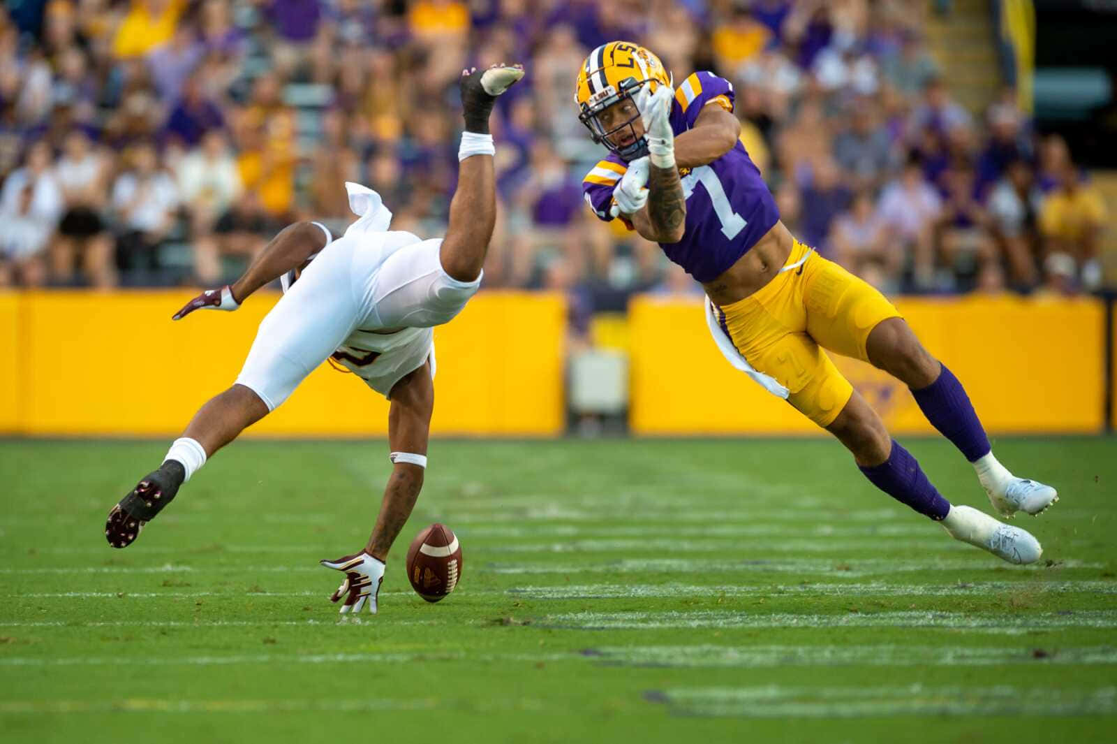 Lsu Football 桌布 壁紙 — 一名身穿紫色和金色球衣的足球員撲向球，而一名身穿白色的防守球員跳起攔截，在充滿活力的綠色球場上。