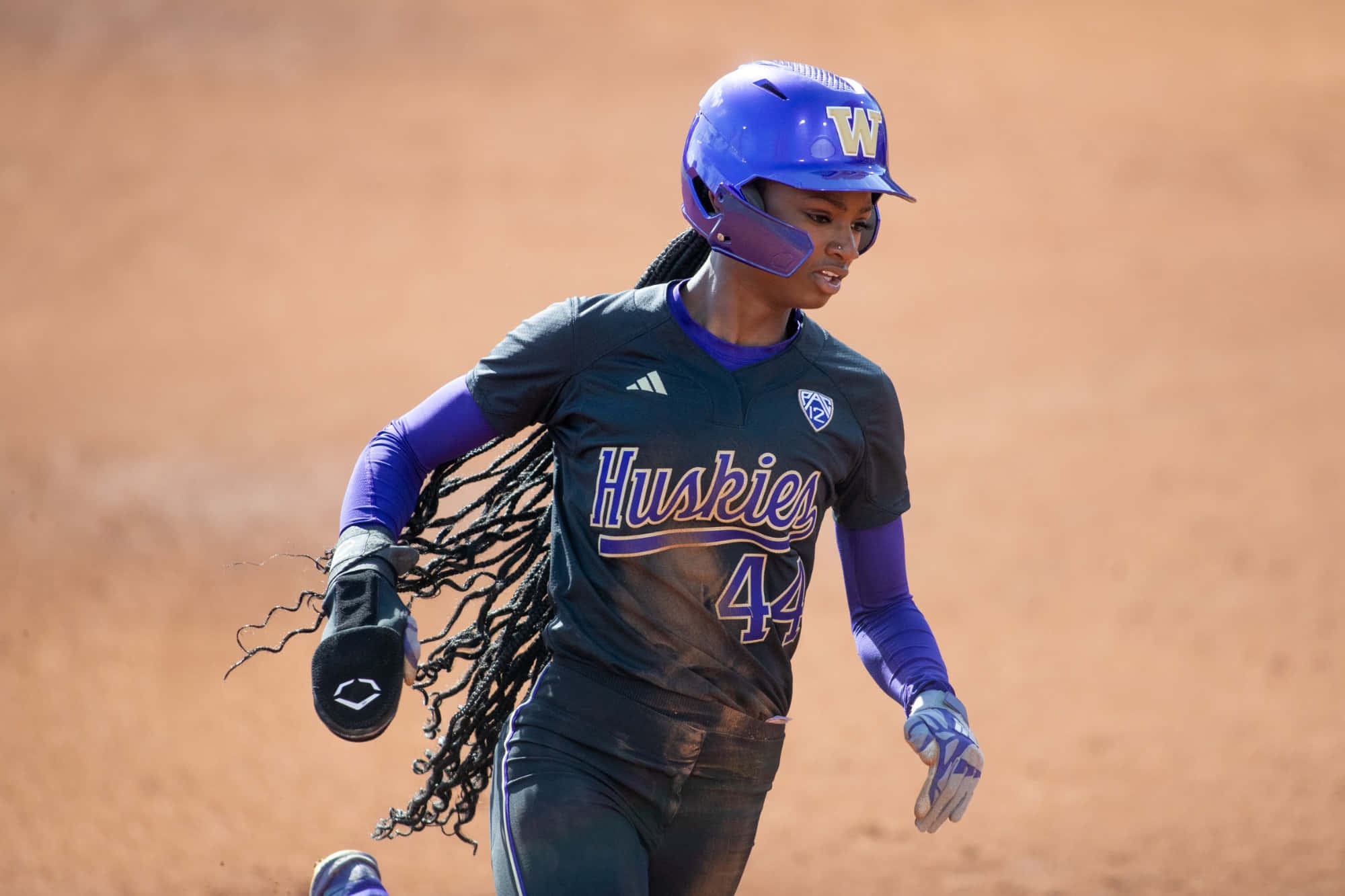 壘球 桌布 壁紙 — 一名身穿紫色和黑色 Huskies 球衣的女壘球運動員在泥地球場上衝刺，辮子飛揚。