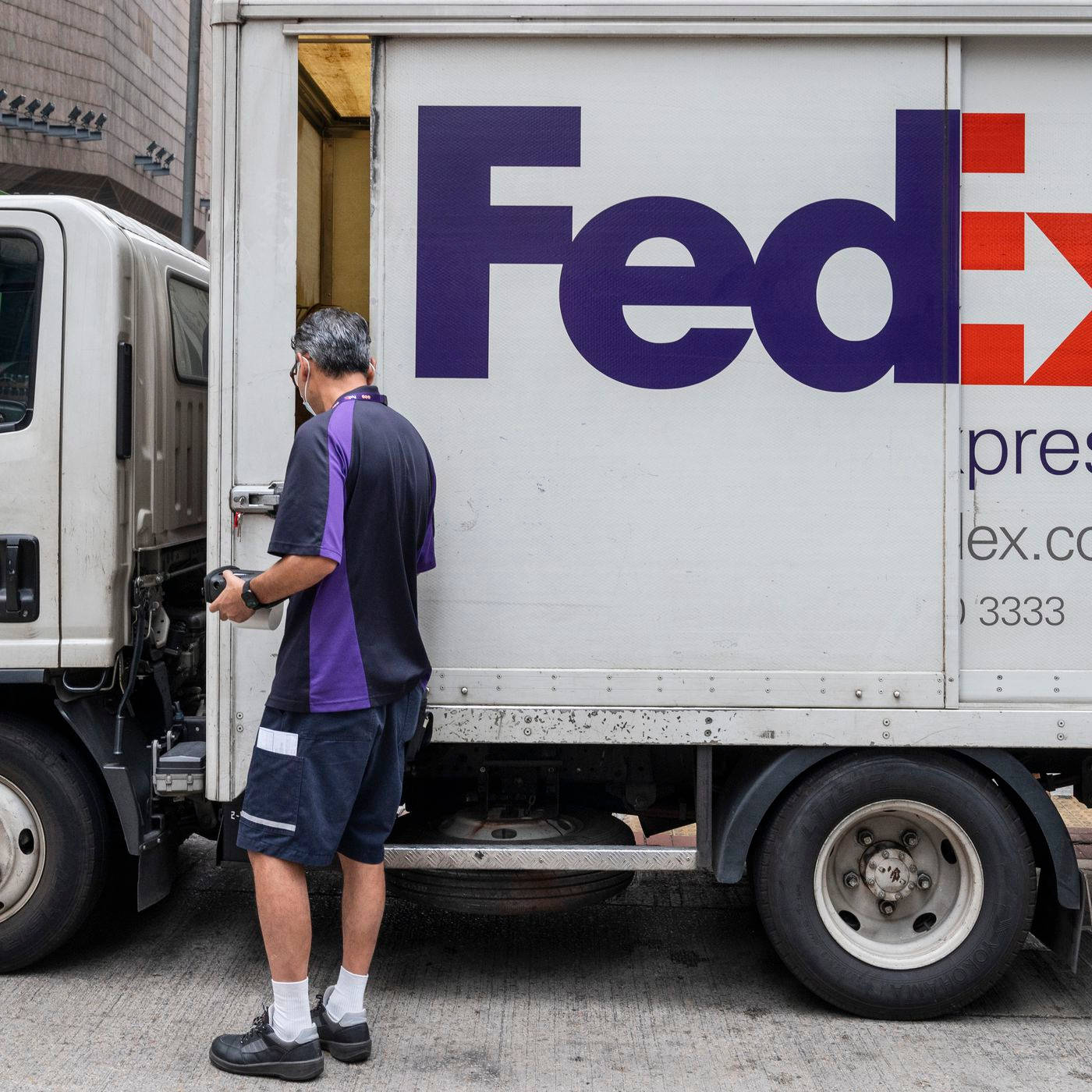 聯邦快遞 桌布 壁紙 — 一名身穿紫色和黑色制服的FedEx送貨司機正在城市街道上將包裹裝載到白色送貨車上。
