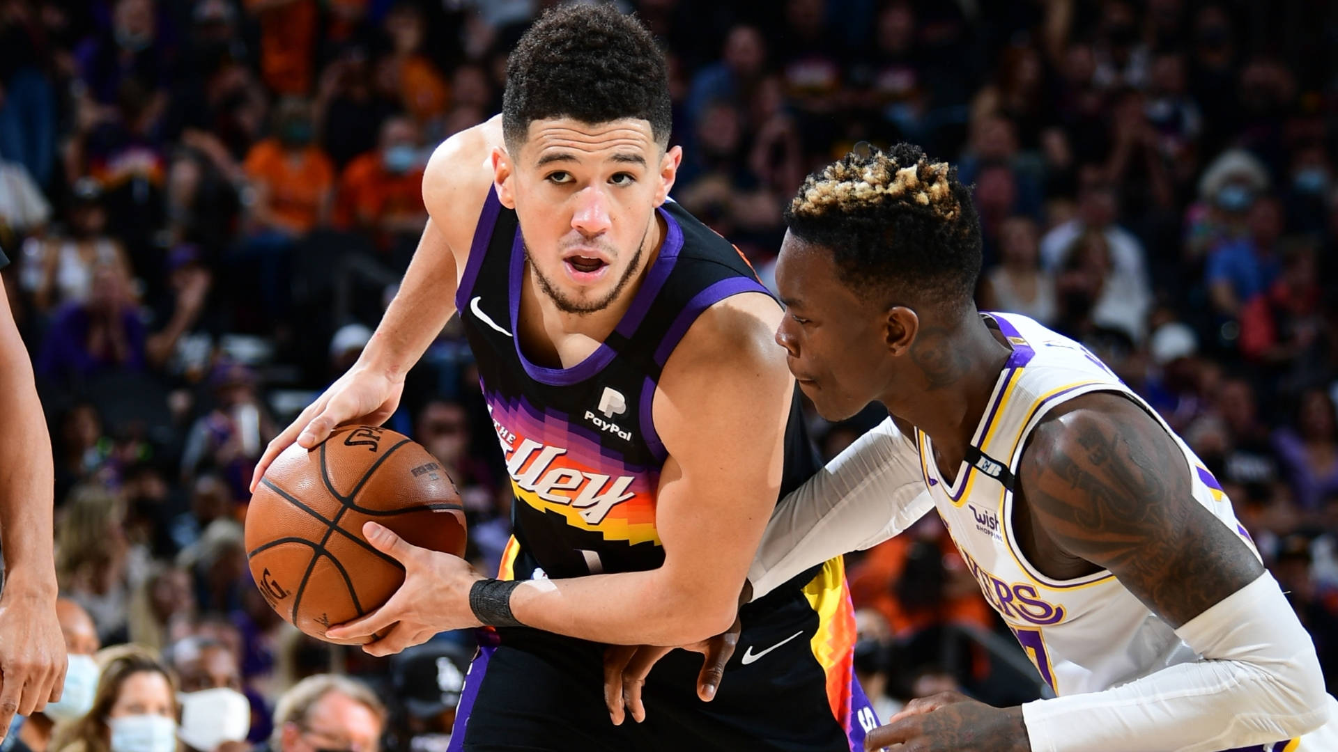 Devin Booker 桌布 壁紙 — 一名身穿紫色和黑色球衣的籃球運動員在擠滿觀眾的場館比賽中運球，同時被一名身穿白色和紫色球衣的對手緊密防守。