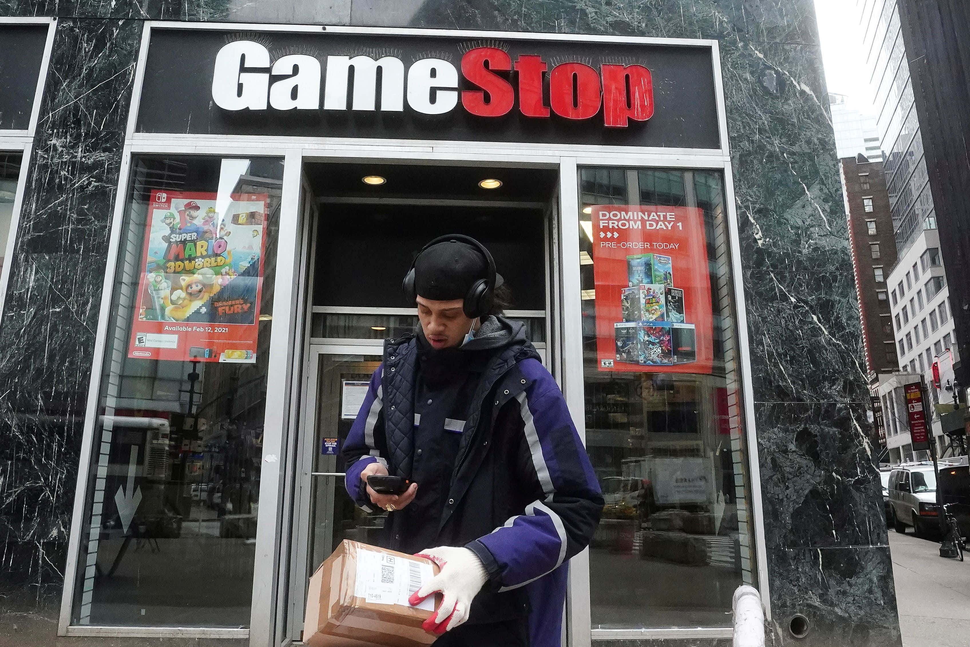 GameStop 桌布 壁紙 — 一名身穿紫色外套、戴著耳機的送貨員站在GameStop店外，手持包裹並查看手機。
