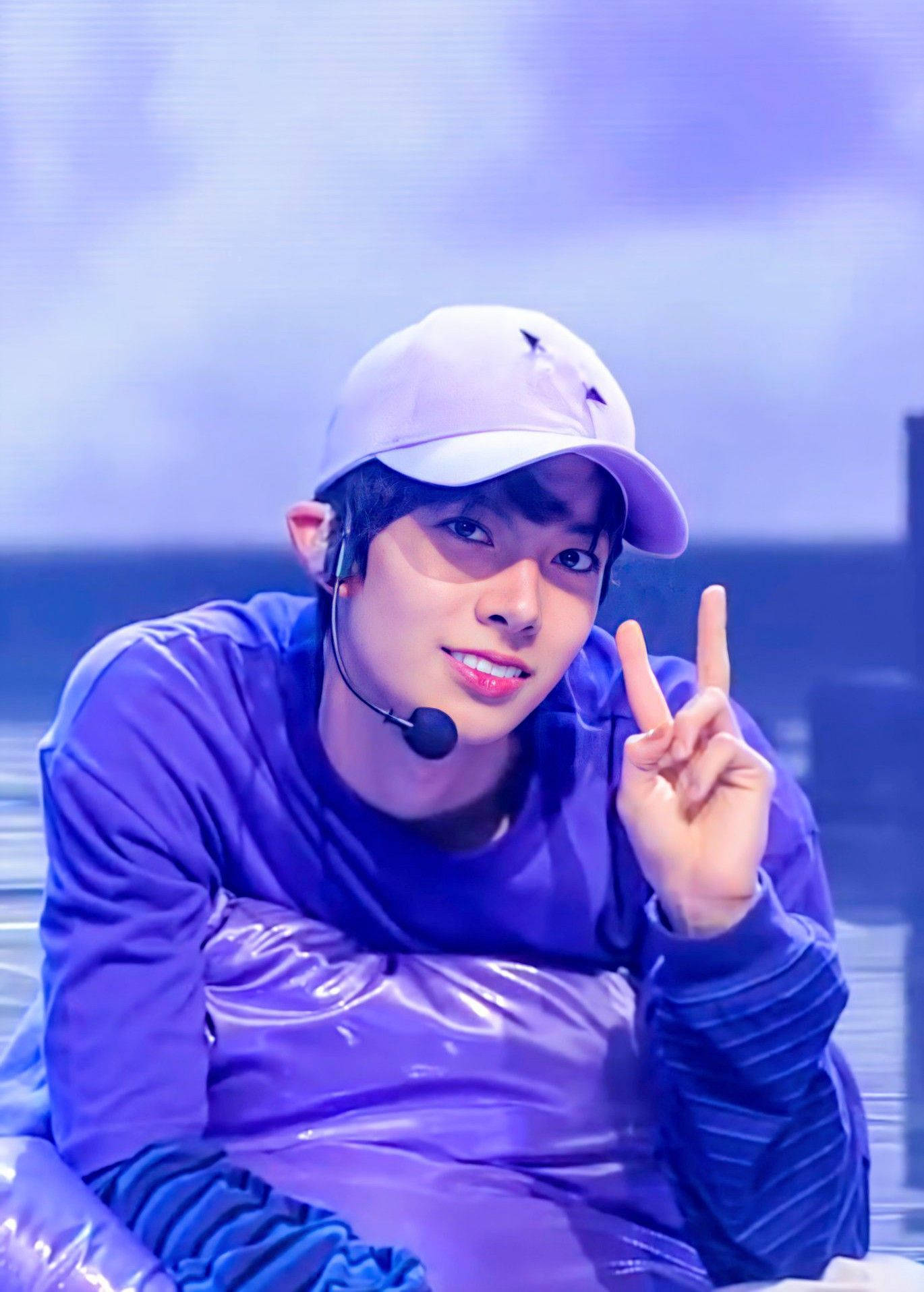 Kpop Aesthetic 桌布 壁紙 — 一名身穿紫色服裝和帽子的年輕男子在藍色舞台燈光下燦爛微笑，同時做出和平手勢。