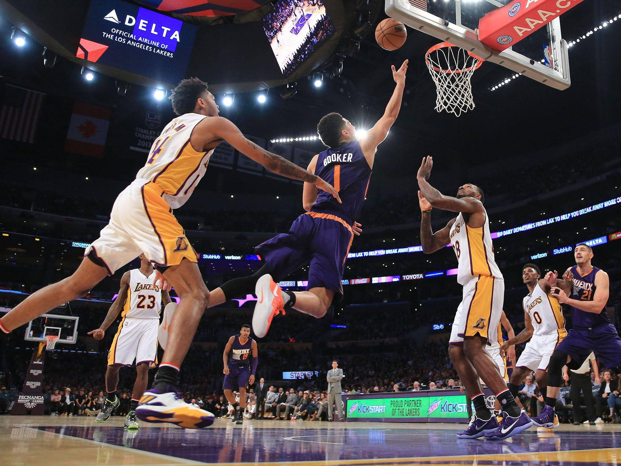 Devin Booker 桌布 壁紙 — 一名身穿紫色球衣的籃球運動員在明亮的場館燈光下跳向籃框，而身穿白色球衣的防守球員試圖封阻他的投籃。