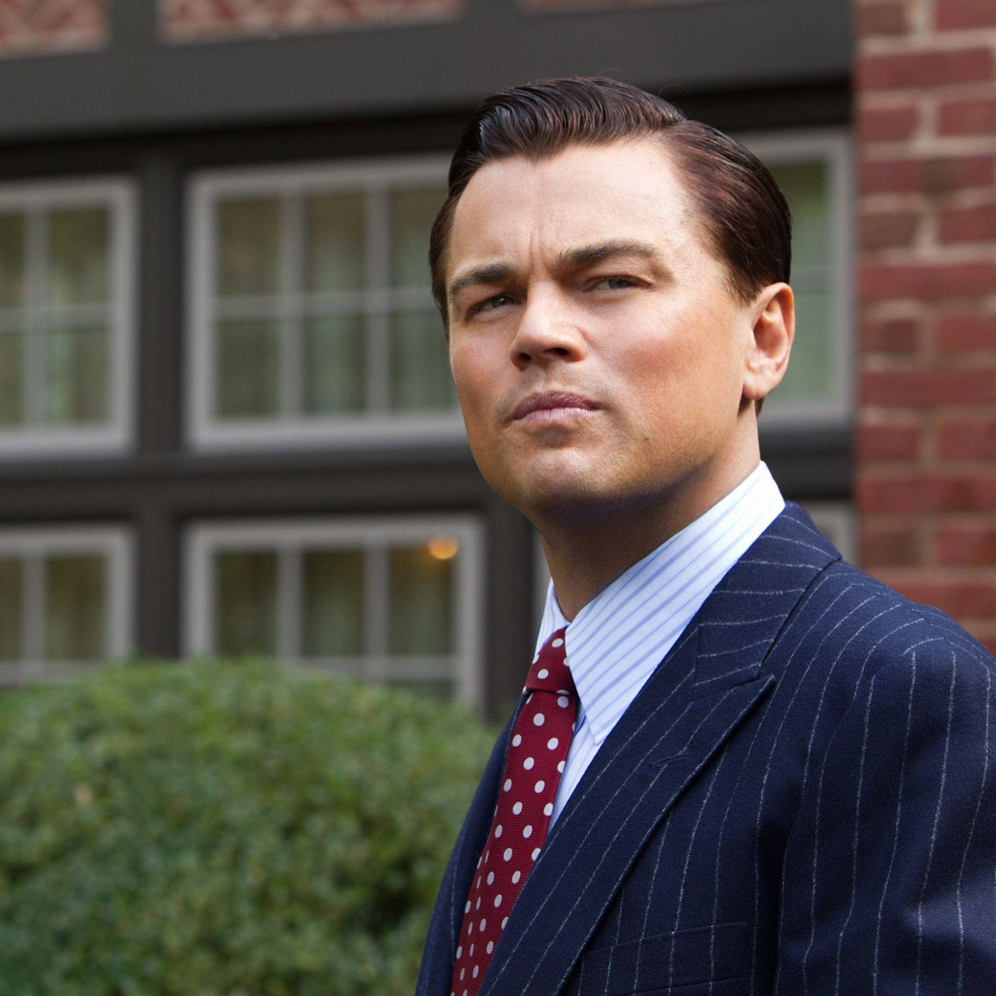 Wolf Of Wall Street 桌布 壁紙 — 一名身穿細條紋西裝、繫著紅色圓點領帶的男子，自信地站在一棟有綠色灌木叢的磚砌建築前。